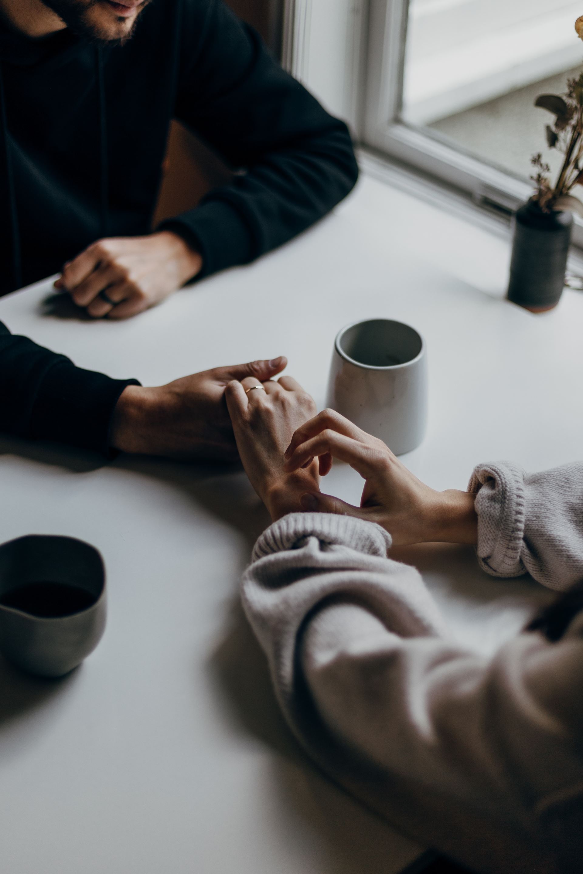 Two people drinking from mugs and holding hands over a white table